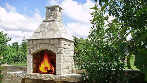 A stone-built outdoor pizza oven with a fire burning inside, surrounded by greenery. - Holiday apartment with air conditioning (Maružini)