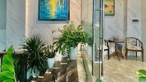 Un vestíbulo con suelo de mármol, una pared de vidrio y dos cuadros. Hay plantas en macetas y una pequeña mesa con sillas.