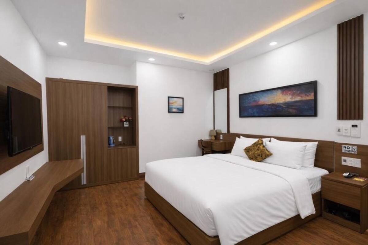 A modern hotel room with a large bed, a flat-screen TV, a wooden wardrobe, a painting on the wall, and a 'No Smoking' sign.
