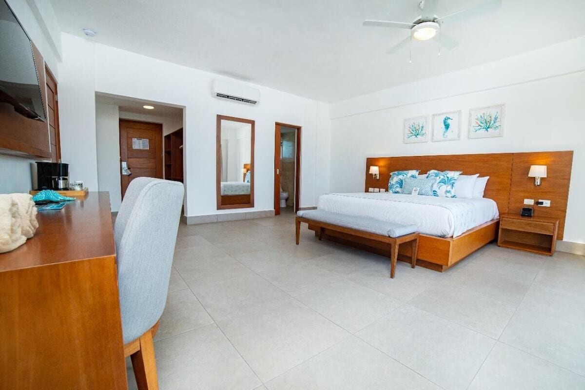 A hotel room with a wooden bed, a desk with a chair, a ceiling fan, and artwork on the wall.