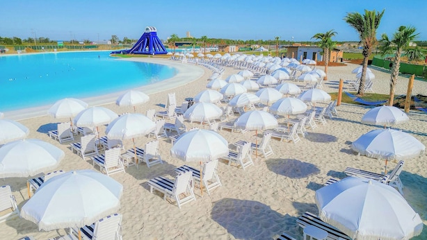 Een strand met vele witte parasols en ligbedden, een helder blauw zwembad en een glijbanenconstructie.