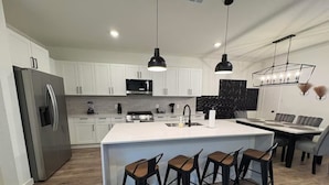 Een moderne keuken met witte kasten, een roestvrijstalen koelkast, een centraal kookeiland met barkrukken en hanglampen.