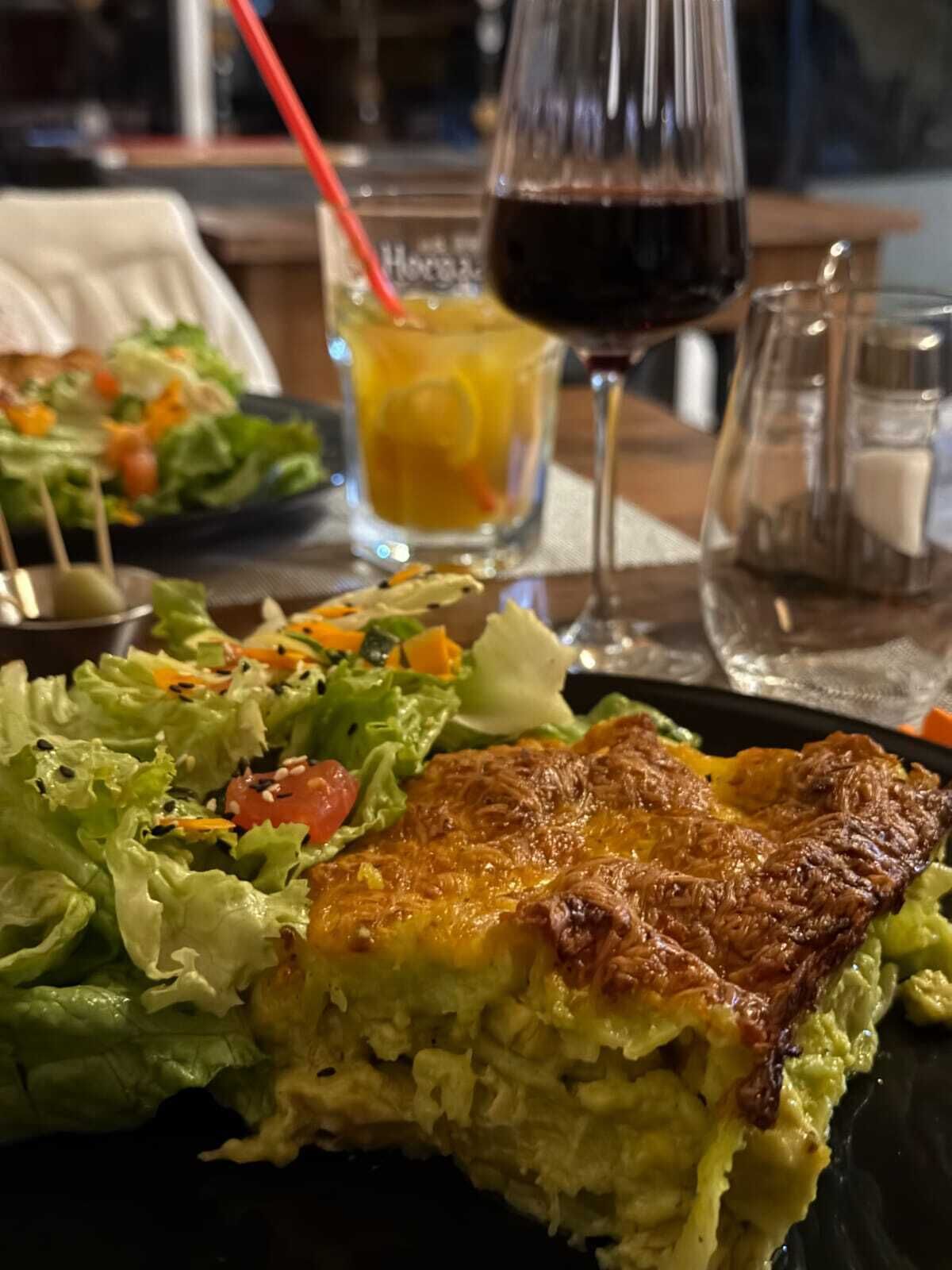 Un plato con una porción de lasaña, una copa de vino tinto y una ensalada sobre una mesa.