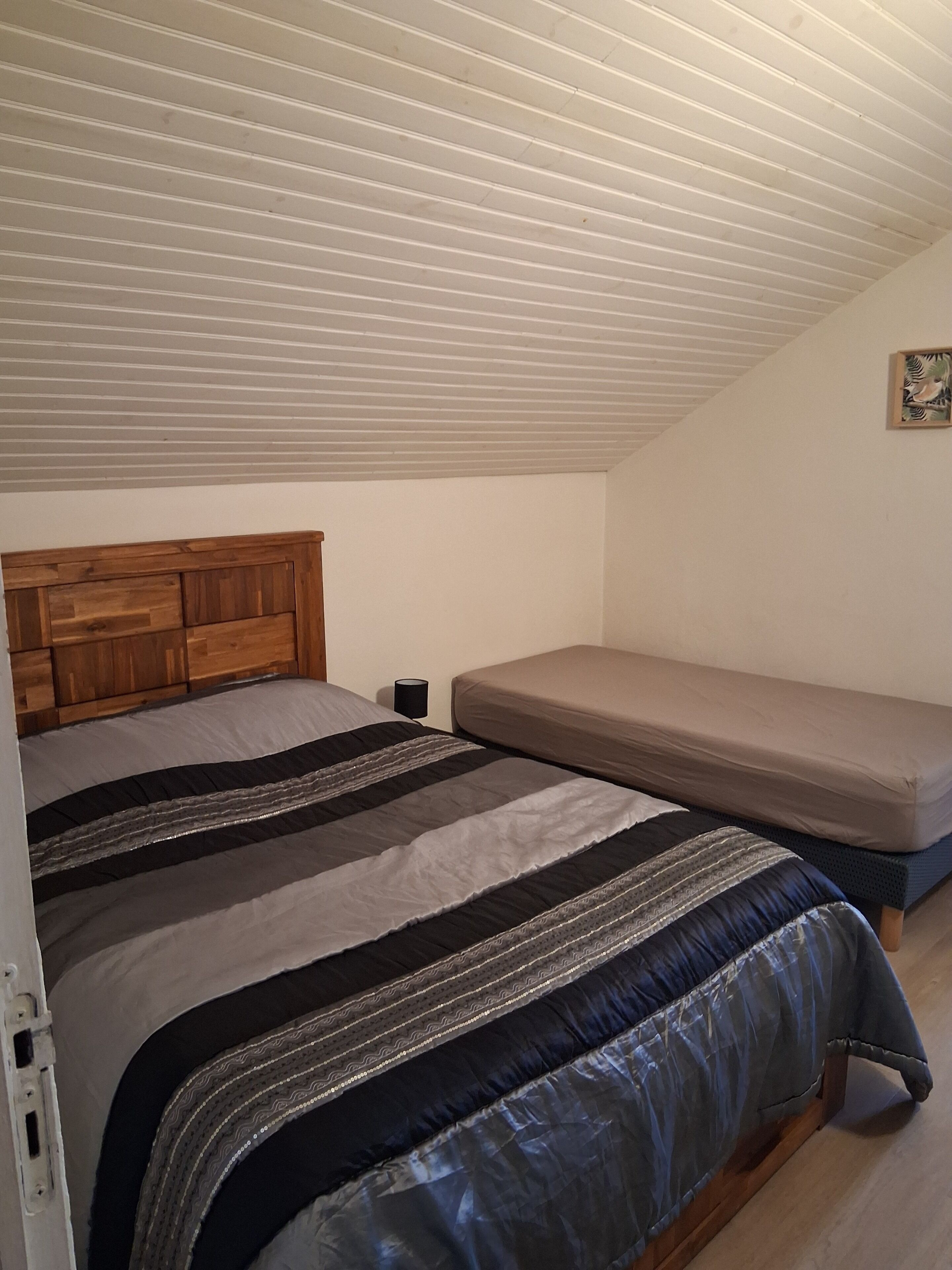 Ein Schlafzimmer mit geneigter Decke, einem Holzkopfteil, einem Bett mit gestreifter Bettwäsche, einem Nachttisch und einem an der Wand hängenden Bild.