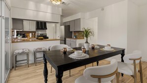 Une cuisine moderne avec des meubles blancs, une table à manger en bois foncé et un luminaire suspendu.