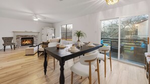 Un salon spacieux doté d’une cheminée, de grandes fenêtres et d’un coin repas avec une table en bois foncé et des chaises blanches.