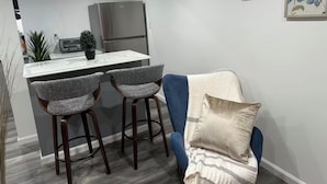 A compact kitchen with a refrigerator, two bar stools, and a blue chair with a cushion. - Cozy Home. (Hempstead)