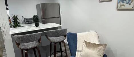 Cocina compacta con refrigerador, dos taburetes de bar y una silla azul con cojín.