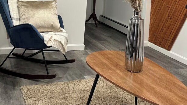 A modern living room with a blue rocking chair, a wooden coffee table, and a vase with dried plants. There are two framed artworks on the wall and a wooden accent wall. - Cozy Home. (Hempstead)