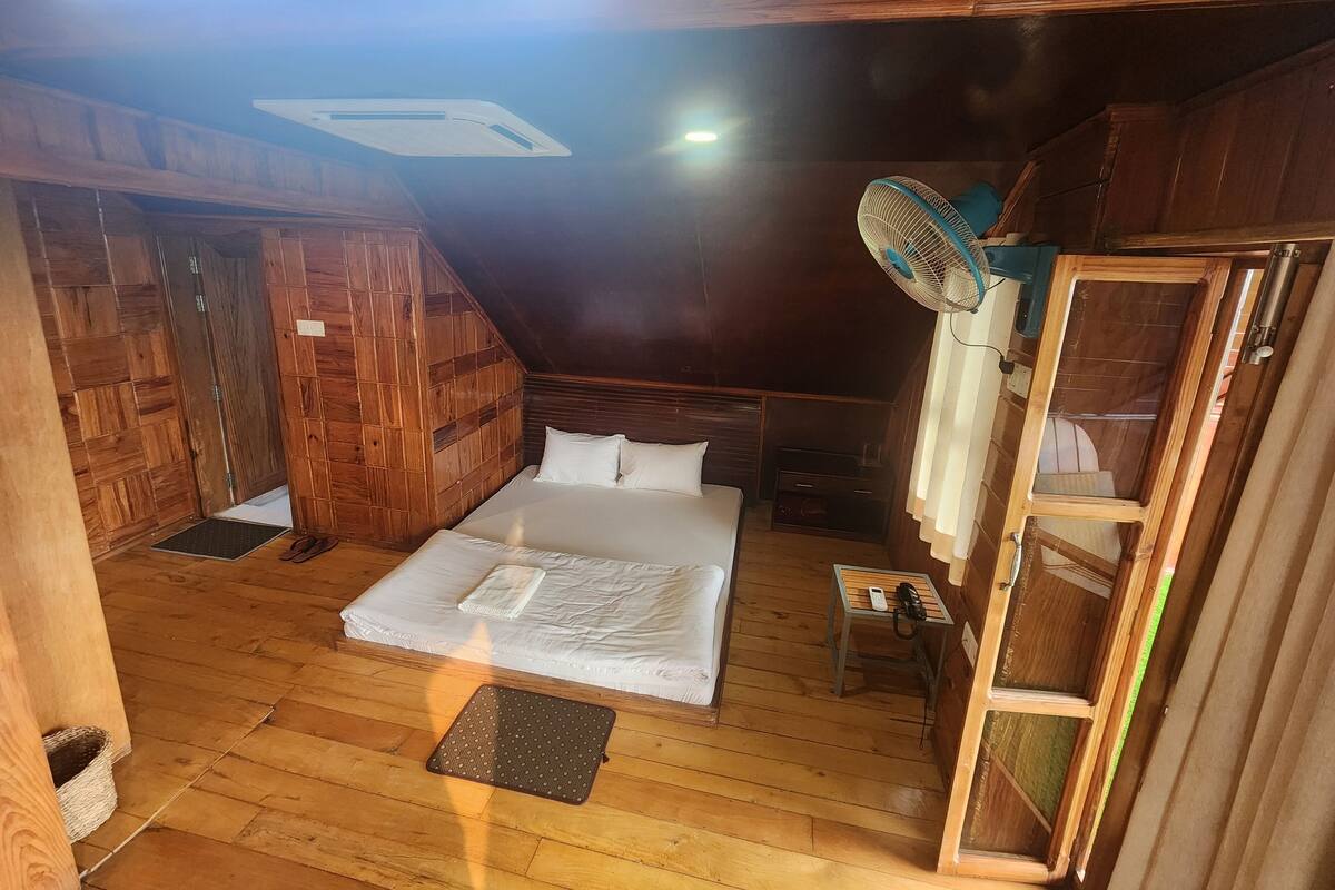 Ein Zimmer mit Holzboden, einem Bett, einem kleinen Tisch mit Telefon und einem Ventilator.