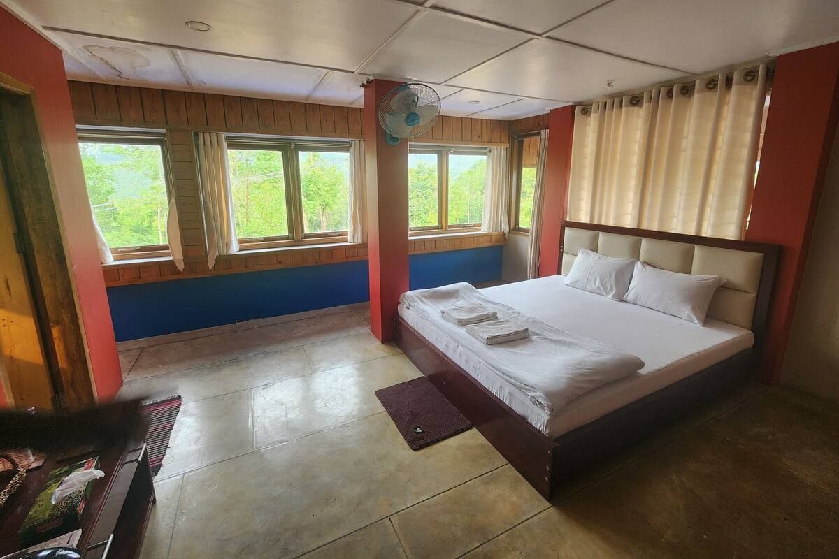 Ein Schlafzimmer mit Bett, Fenstern, Deckenventilator und Blick auf Grünfläche.