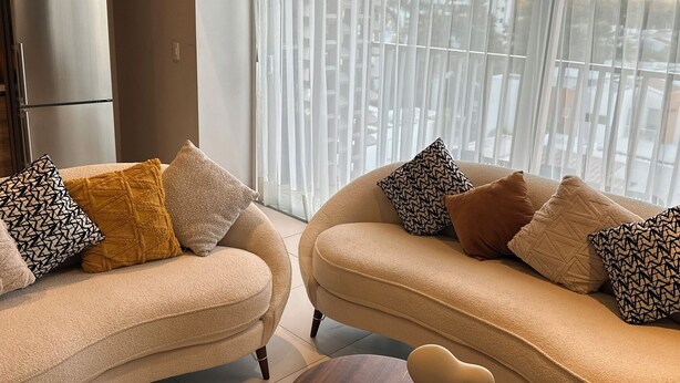 Un soggiorno moderno con due divani beige, un tavolino in legno e vista sulla città attraverso tende leggere.