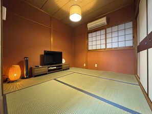 Ein traditioneller japanischer Raum mit Tatami-Boden, einem Fernseher auf einem Holzbodenständer, einer Wand-Klimaanlage und einem Shoji-Bildschirm.
