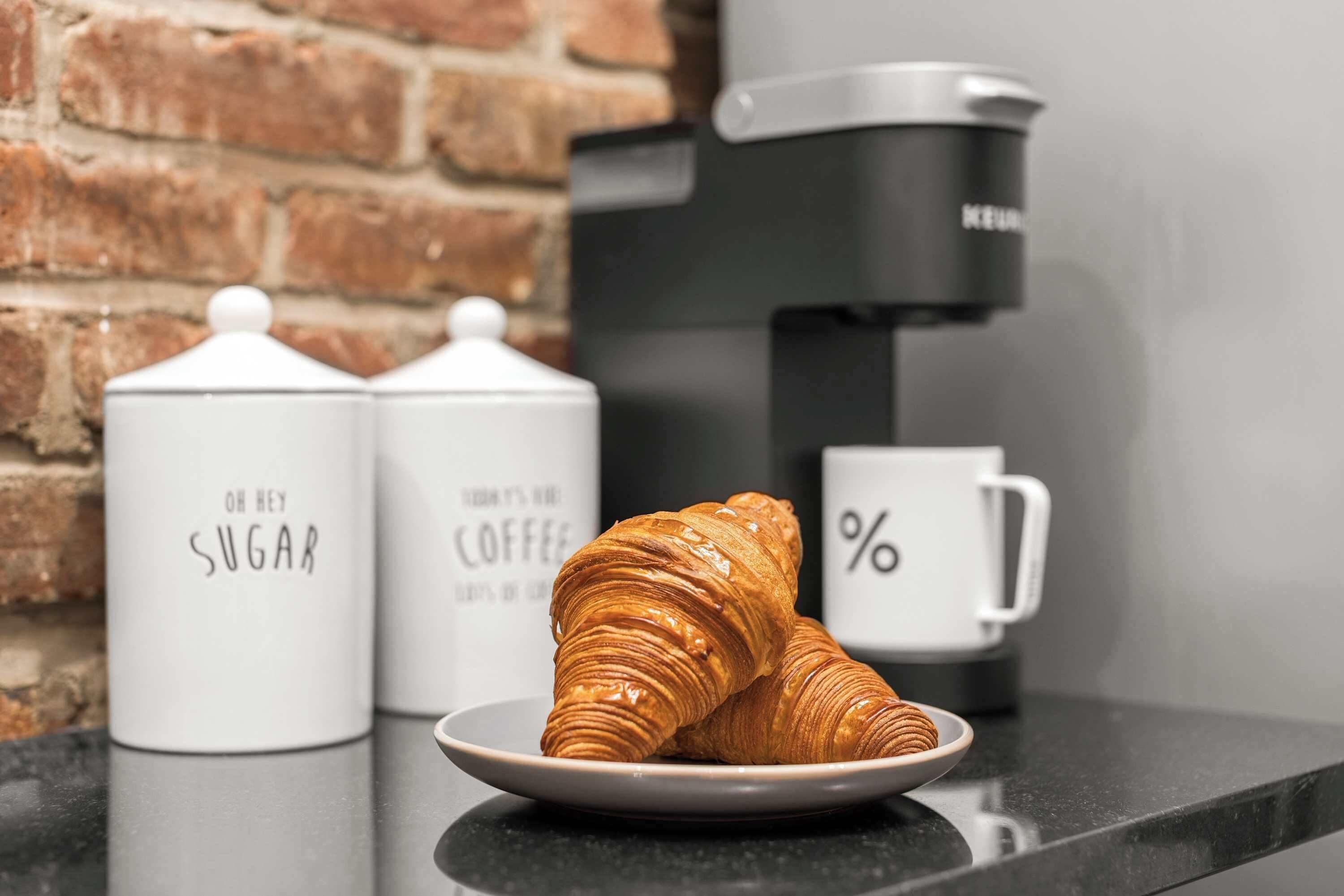 Ein Croissant auf einem Teller, eine Kaffeemaschine und zwei Zuckerbehälter auf einer Küchenarbeitsplatte.