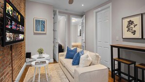 A living room with a brick wall, a ceiling fan, a flat-screen TV mounted on the wall, a white sofa with blue and white cushions, a small white table with a plant, and a wooden coffee table. - Spacious Midtown Retreat 2BR NYC Haven (New York)