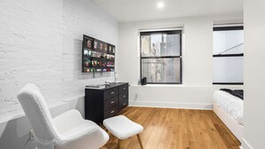 A modern bedroom with a white brick wall, wooden flooring, a black dresser, a white chair, a footstool, a bed, and a window with a view of buildings. - Prime NYC Location 4BR Spacious Haven (New York)