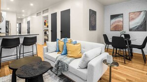 A modern living room with a white sofa, black chairs, a round dining table, and a kitchen in the background. - Prime NYC Location 4BR Spacious Haven (New York)