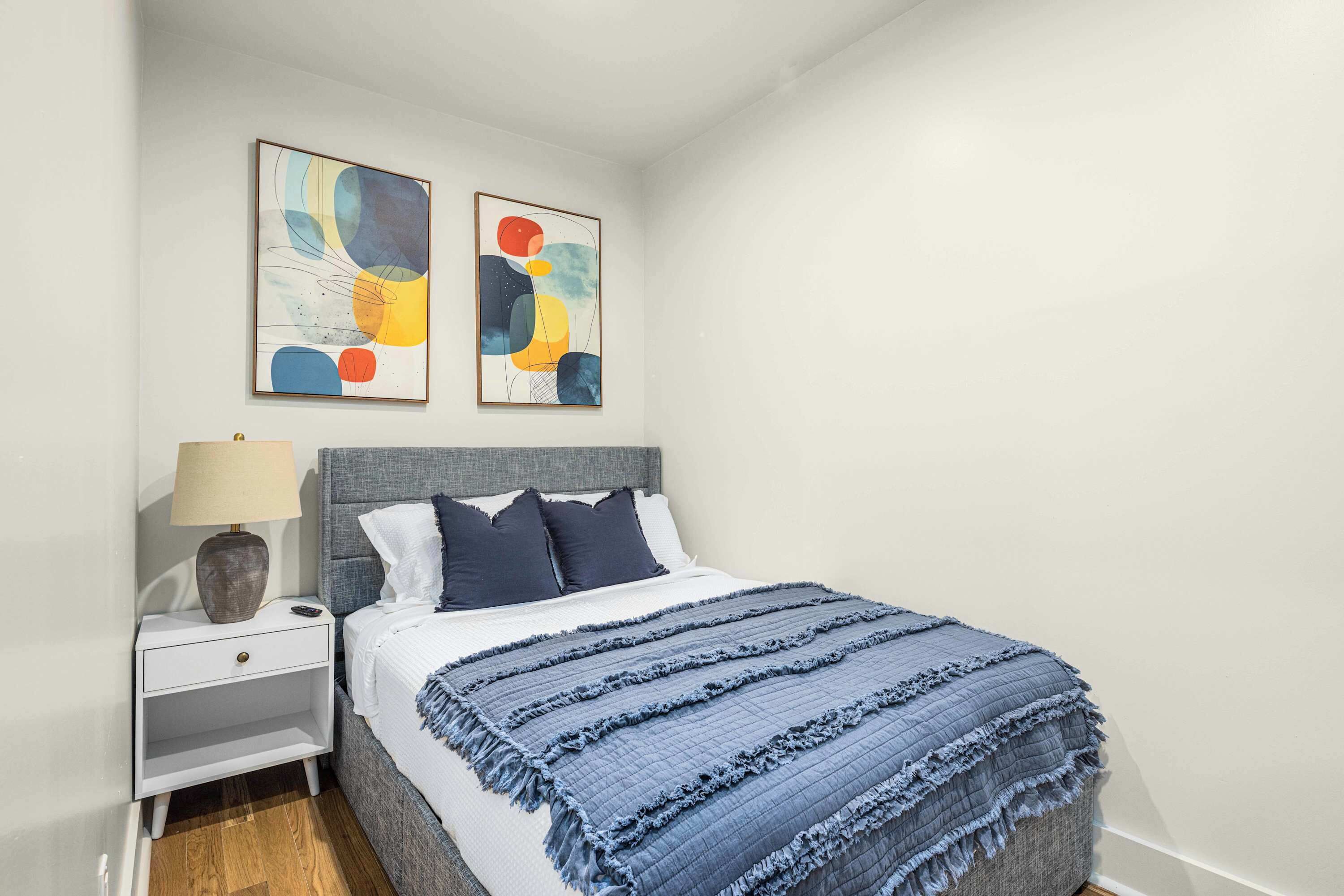 Un dormitorio con cama, mesita de noche, lámpara y pinturas abstractas en la pared.