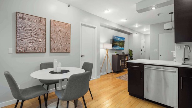 Una cocina moderna con armarios oscuros, encimera blanca y lavavajillas empotrado. Hay una zona de comedor con una mesa redonda y sillas grises. Dos cuadros abstractos decoran la pared.
