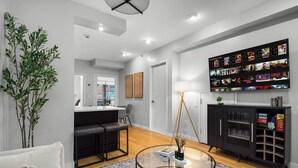 Una sala moderna con una mesa de centro de vidrio, un sofá negro, una zona de comedor con una mesa blanca y un televisor montado en la pared mostrando opciones de películas.