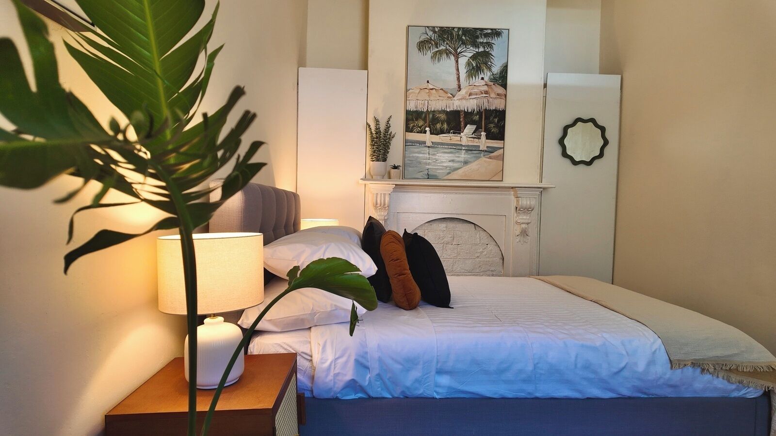 A hotel room with a bed, a nightstand with a lamp, a large plant, a fireplace, and a painting of a tropical scene.