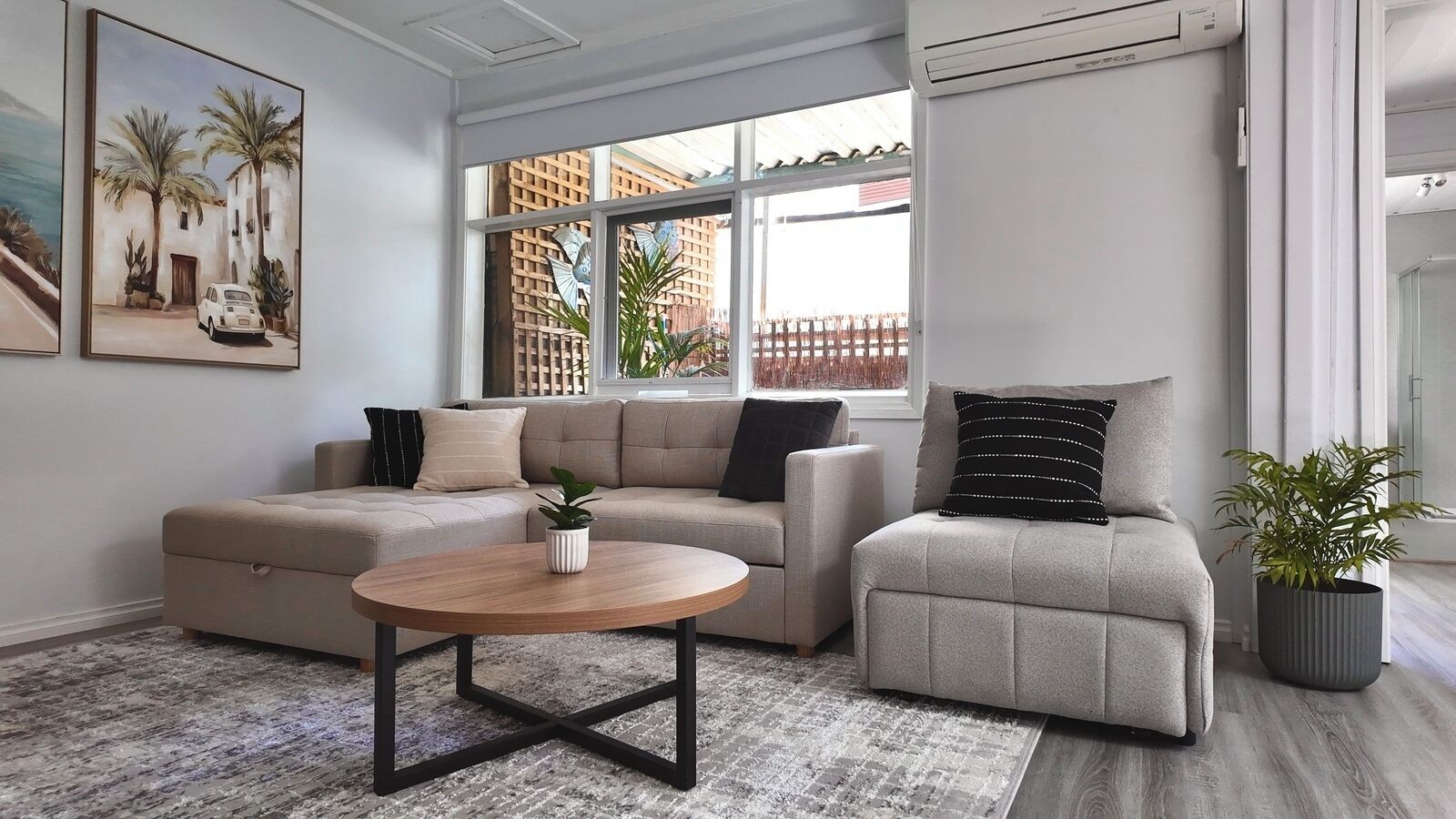 A modern living room with a beige sectional sofa, a round coffee table, and a grey armchair. There is a large window with a view of palm trees and a fence.
