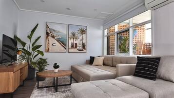 A modern living room with a sofa, a coffee table, and a TV on a wooden cabinet. There are two large windows providing natural light and a view of buildings outside.