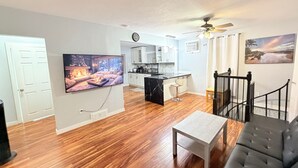 A spacious living room with hardwood flooring, a large flat-screen TV, a fireplace, a kitchen with white cabinets, and a dining area with a bar stool.
