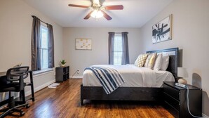 A bedroom with a bed, a desk, a chair, a ceiling fan, and a window with curtains.