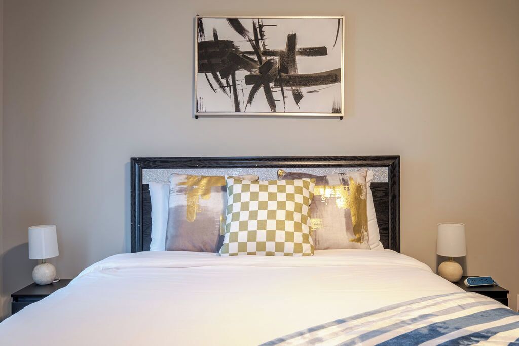 Een net opgemaakt bed met een gestreepte dekbedovertrek, een ruitkussen en een lijst met een abstract schilderij erboven.