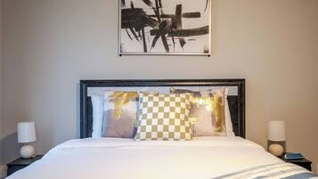 Een net opgemaakt bed met een gestreepte dekbedovertrek, een ruitkussen en een lijst met een abstract schilderij erboven.