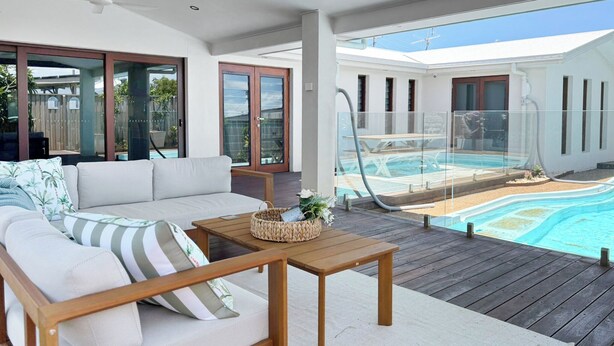 Une terrasse en bois avec un canapé, une table et une piscine visible à travers des portes vitrées.
