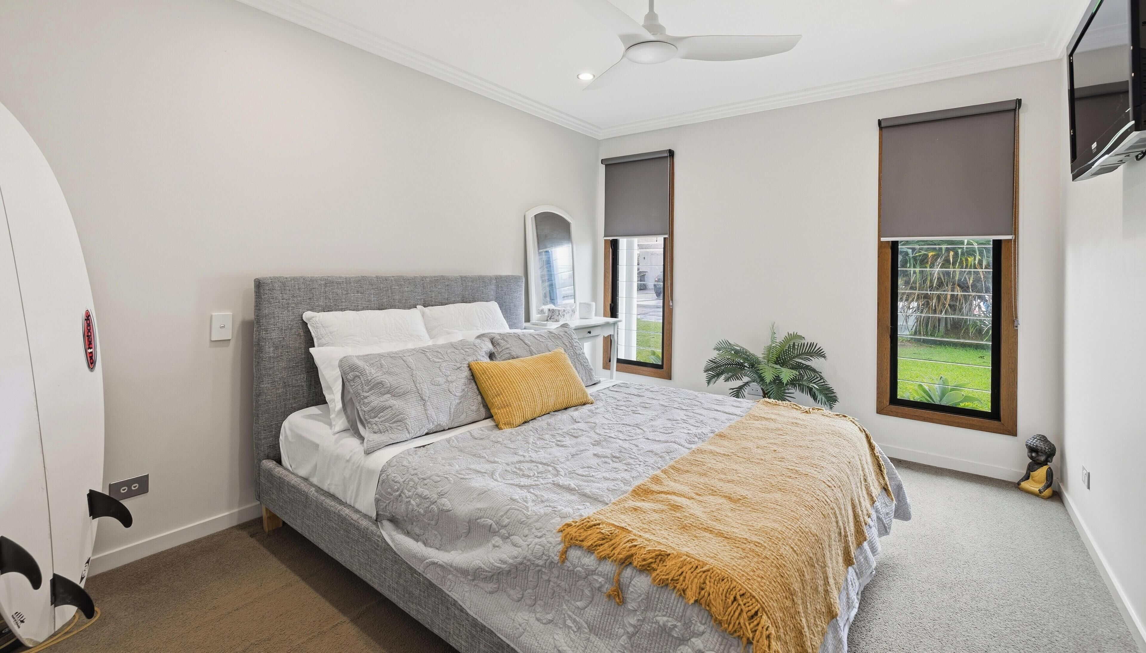 Un dormitorio con cama, ventilador de techo, ventana con vistas a follaje y una tabla de surf apoyada contra la pared.