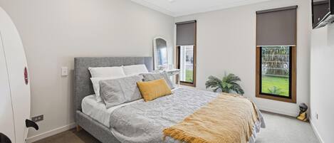 Un dormitorio con cama, ventilador de techo, ventana con vistas a follaje y una tabla de surf apoyada contra la pared.