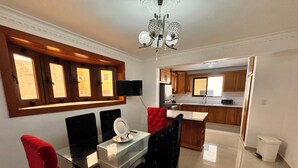 A modern dining area with a glass table, red and black upholstered chairs, a chandelier, and a kitchen in the background. - Enjoy Huge 4 Bedrooms and 3 Bathrooms (Santiago de los Caballeros)