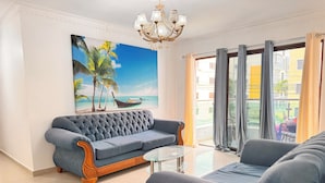 A living room with a blue sofa, a glass coffee table, and a painting of a beach scene. - Enjoy Huge 4 Bedrooms and 3 Bathrooms (Santiago de los Caballeros)