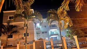 A nighttime view of a residential building with multiple balconies, palm trees, and a gated entrance. - Enjoy Huge 4 Bedrooms and 3 Bathrooms (Santiago de los Caballeros)