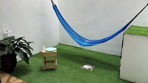 Eine Hängematze, zwischen zwei Wänden in einer Ecke befestigt, mit Kunstgraseinlage, einem kleinen Holzschemel und einem Blumentopf.