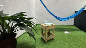 Ein kleiner Holzschemel mit einem Glas darauf, platziert auf künstlichem Rasen, mit einer blauen Hängematte in der Nähe.