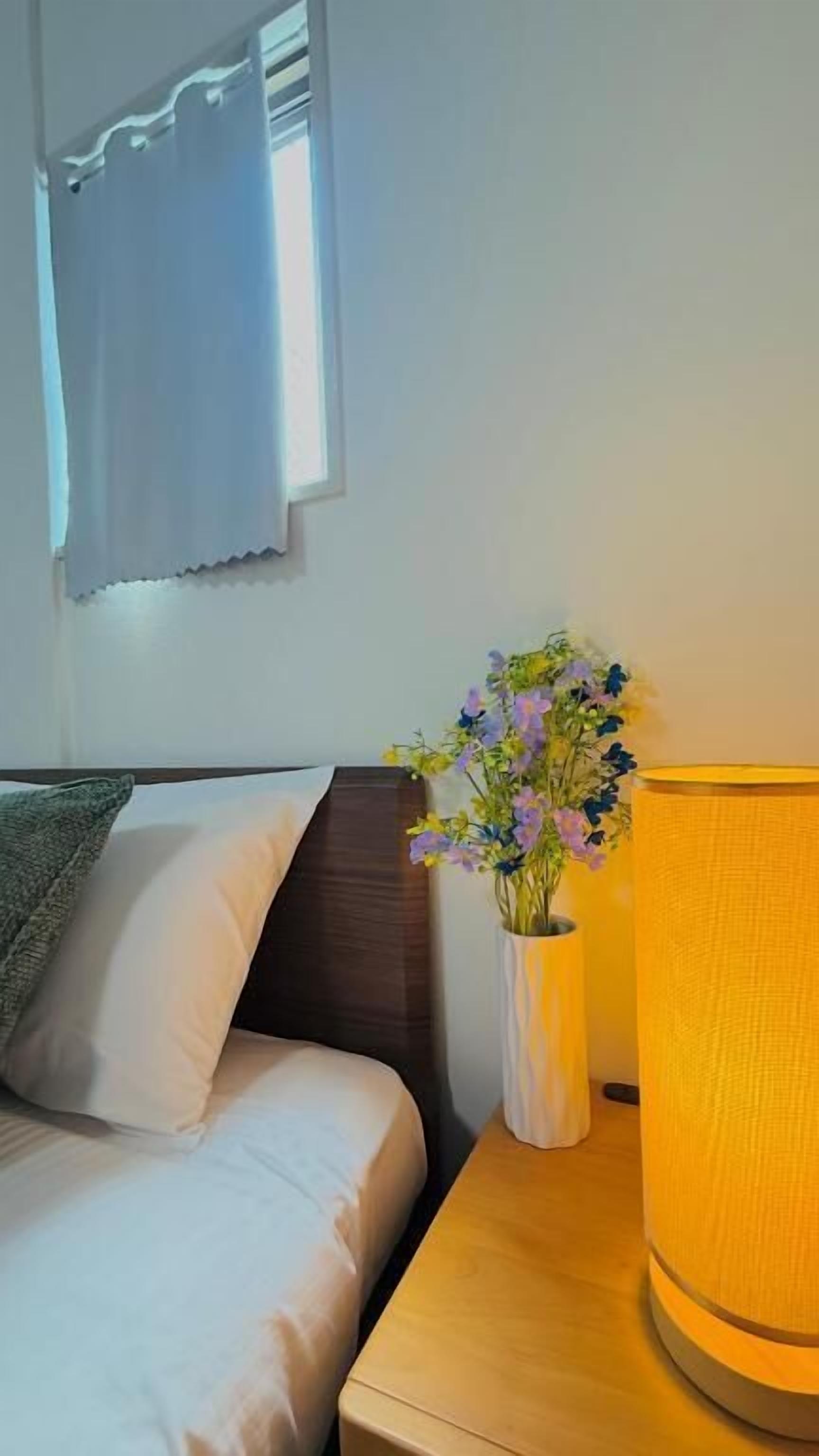 Een net opgemaakt bed met witte lakens, een houten nachtkastje met een lamp en een vaas met bloemen.