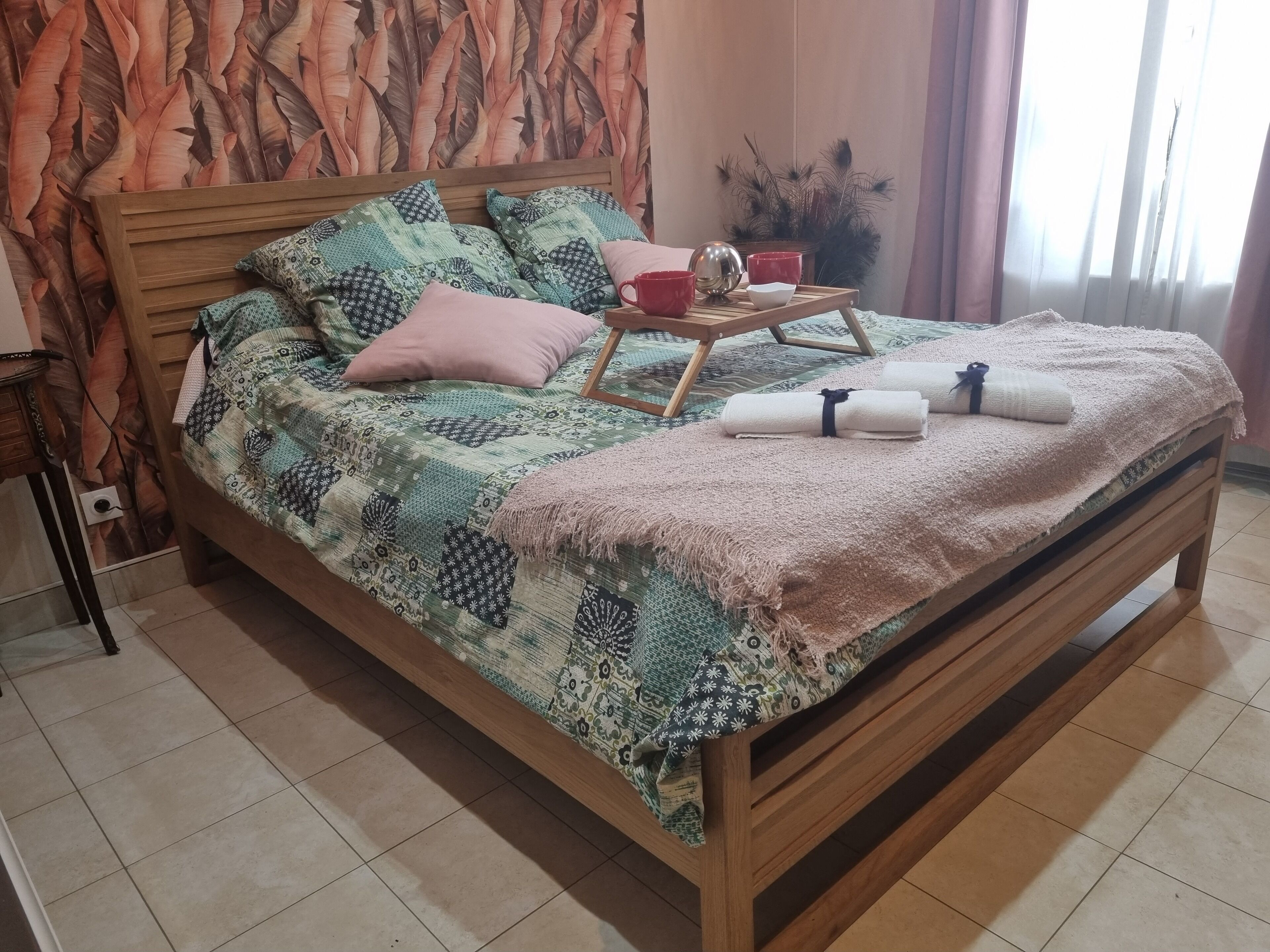 A neatly made bed with a patterned bedspread, pillows, and a bedside table with a tray holding a bowl and a cup.