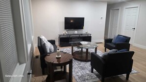 Una sala de estar moderna con un televisor de pantalla plana, sofás negros, una mesa de centro de vidrio y una alfombra estampada.
