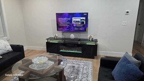 Una sala de estar moderna con un televisor de pantalla plana montado en la pared, un mueble de entretenimiento negro con frentes de vidrio y un teléfono inteligente Galaxy S25 Ultra sobre una mesa de centro de vidrio.