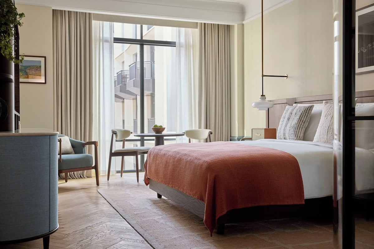 A hotel room with a large bed, a desk with a chair, a small table, and a balcony with a view of the building.