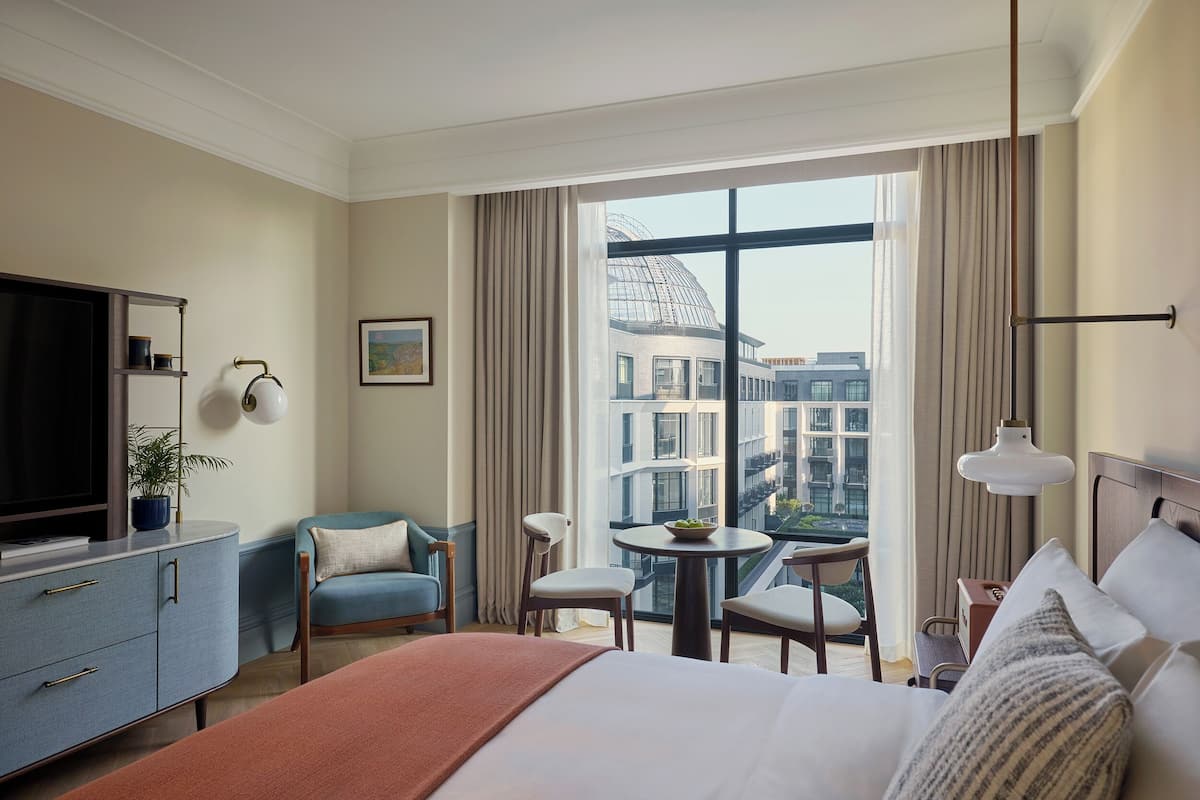 A hotel room with a large bed, a TV, a desk with a chair, a small table with a bowl of fruit, and a view of buildings through large windows.
