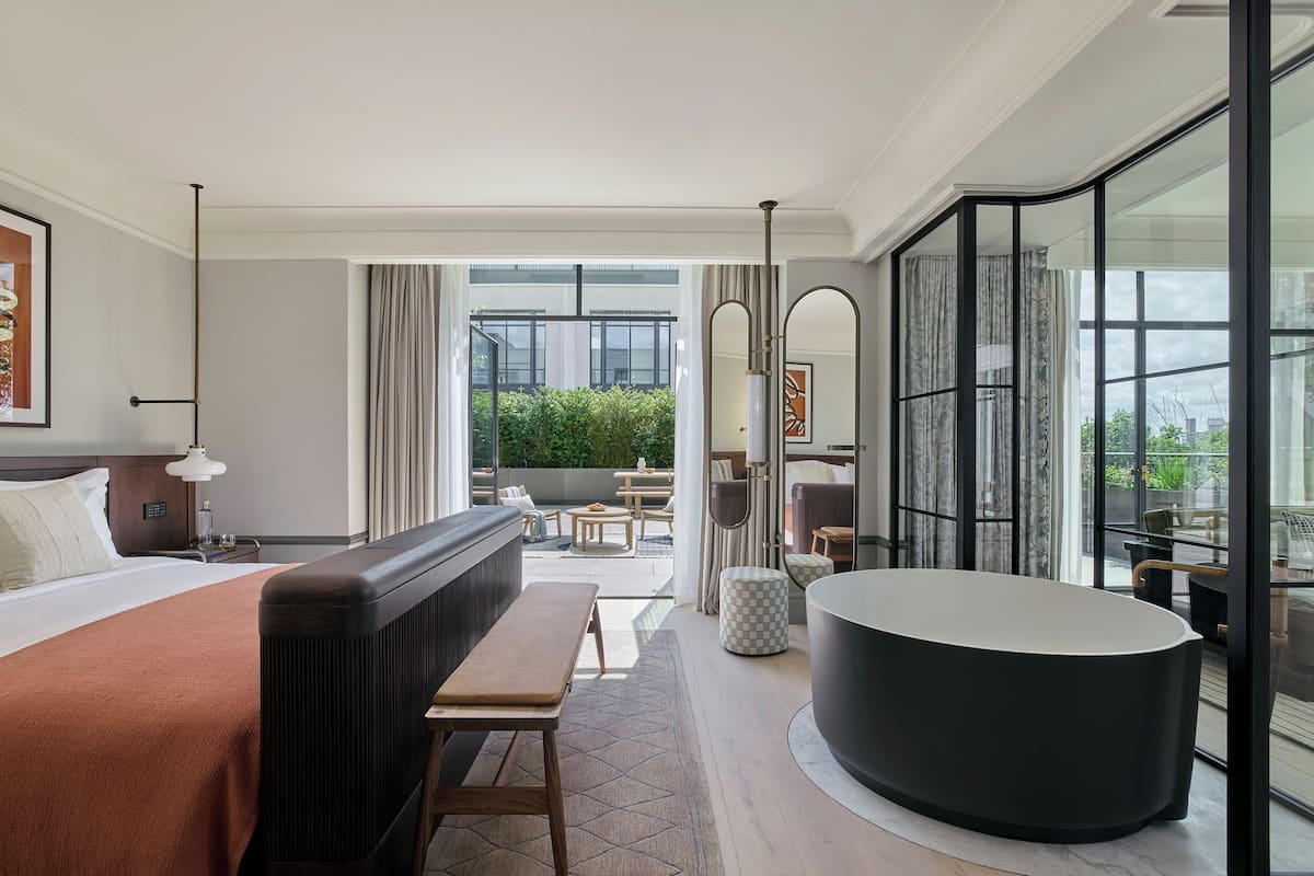 A modern hotel room with a large bed, a sitting area, a bathtub, and a view of the outdoors.