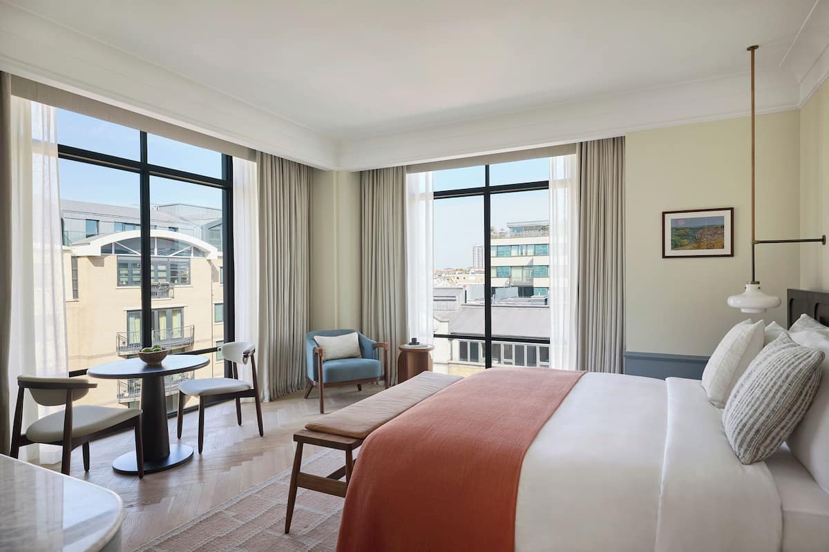 A hotel room with a large bed, a dining table with chairs, a small round table, and a balcony with a chair.