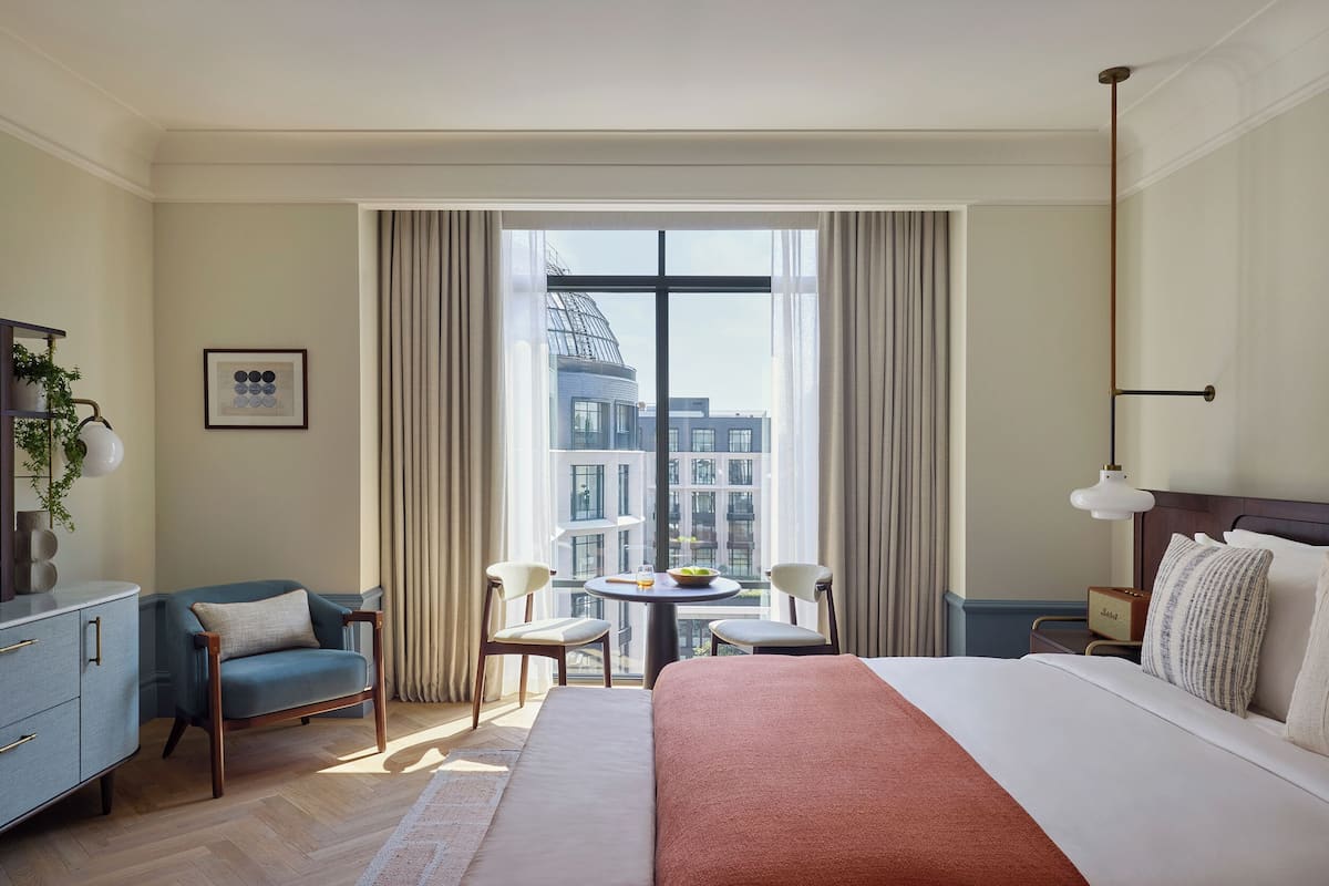 A hotel room with a large bed, a desk with a chair, a small table with two chairs, and a view of the city through large windows.