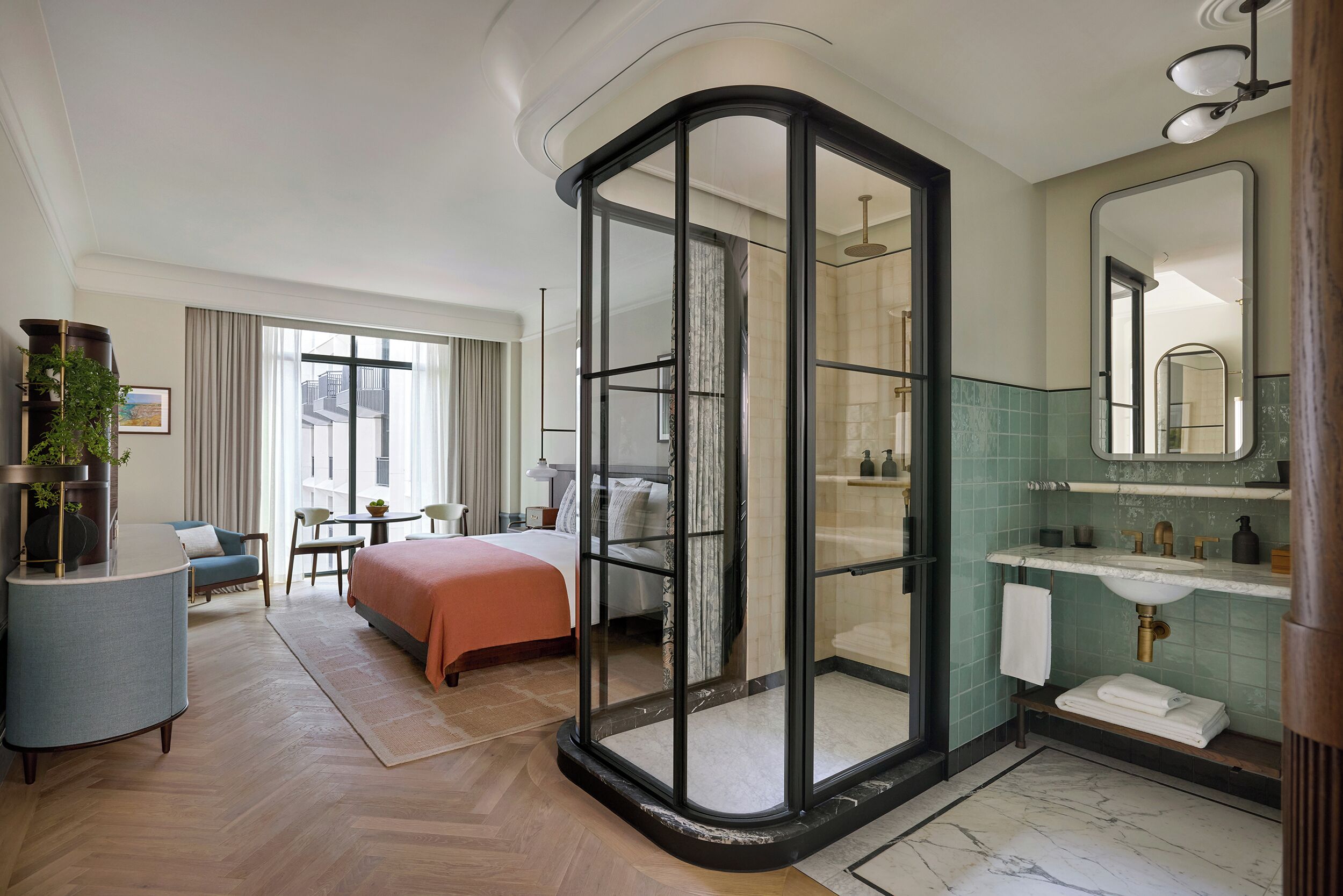 A hotel room with a bed, a desk, a chair, a glass-enclosed shower, a bathroom with a sink and mirror, and a view of the outside.