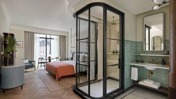 A hotel room with a bed, a desk, a chair, a glass-enclosed shower, a bathroom with a sink and mirror, and a view of the outside.